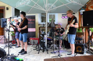 Outdoor band performance under a large umbrella; guitarist and singer at front, drummer in back, all with amplifiers and cables visible.