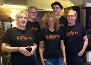 Five people wearing black Garage 66 t-shirts posing together in a casual venue with brick walls in the background.
